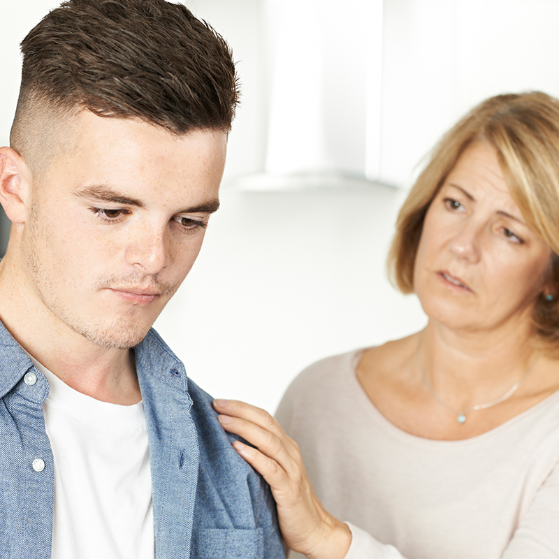 Photo of troubled looking young adult with mom placing reassuring hand on his shoulder.